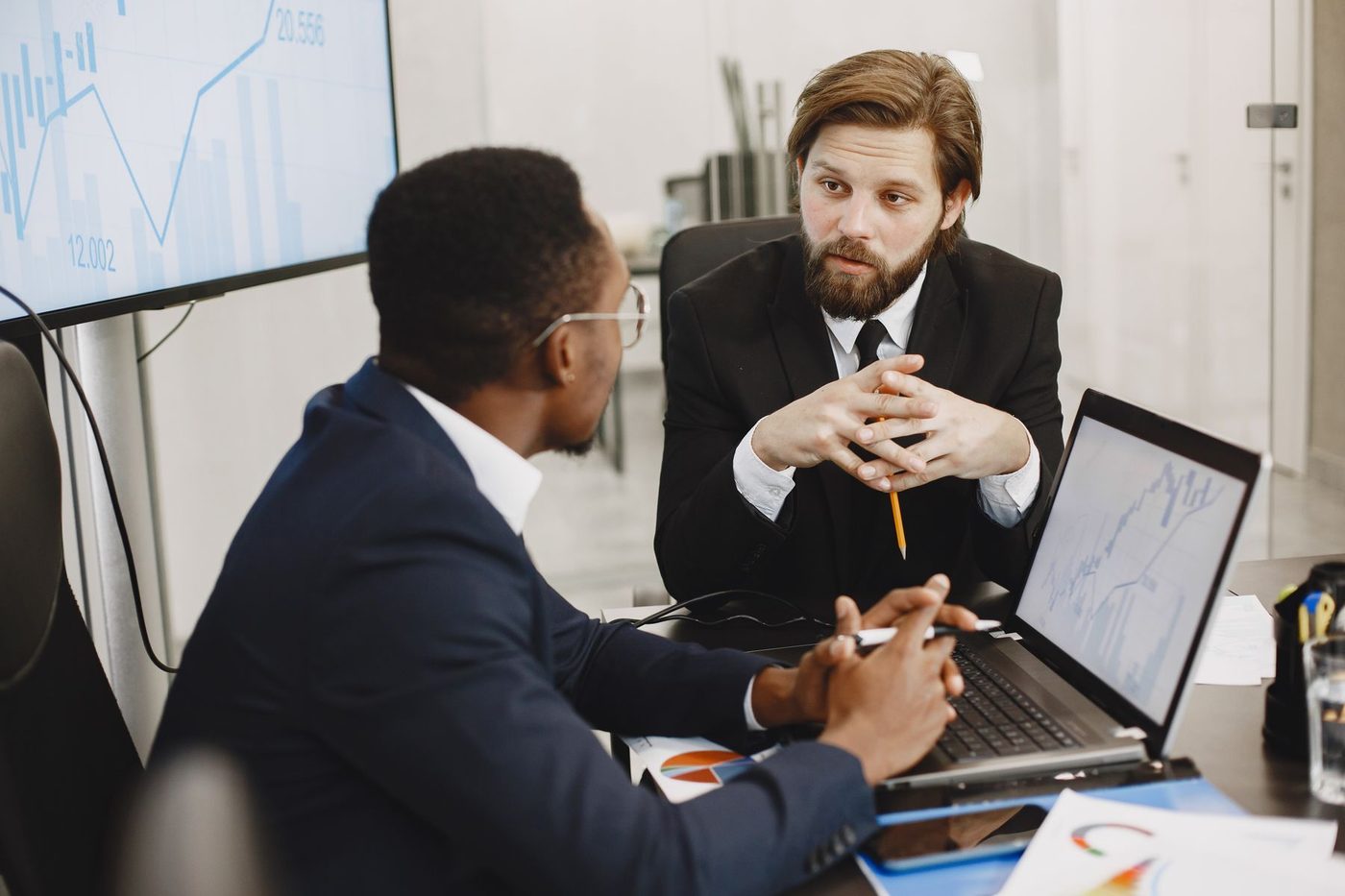 Two business owners at a meeting table, one listening with his hands clasped