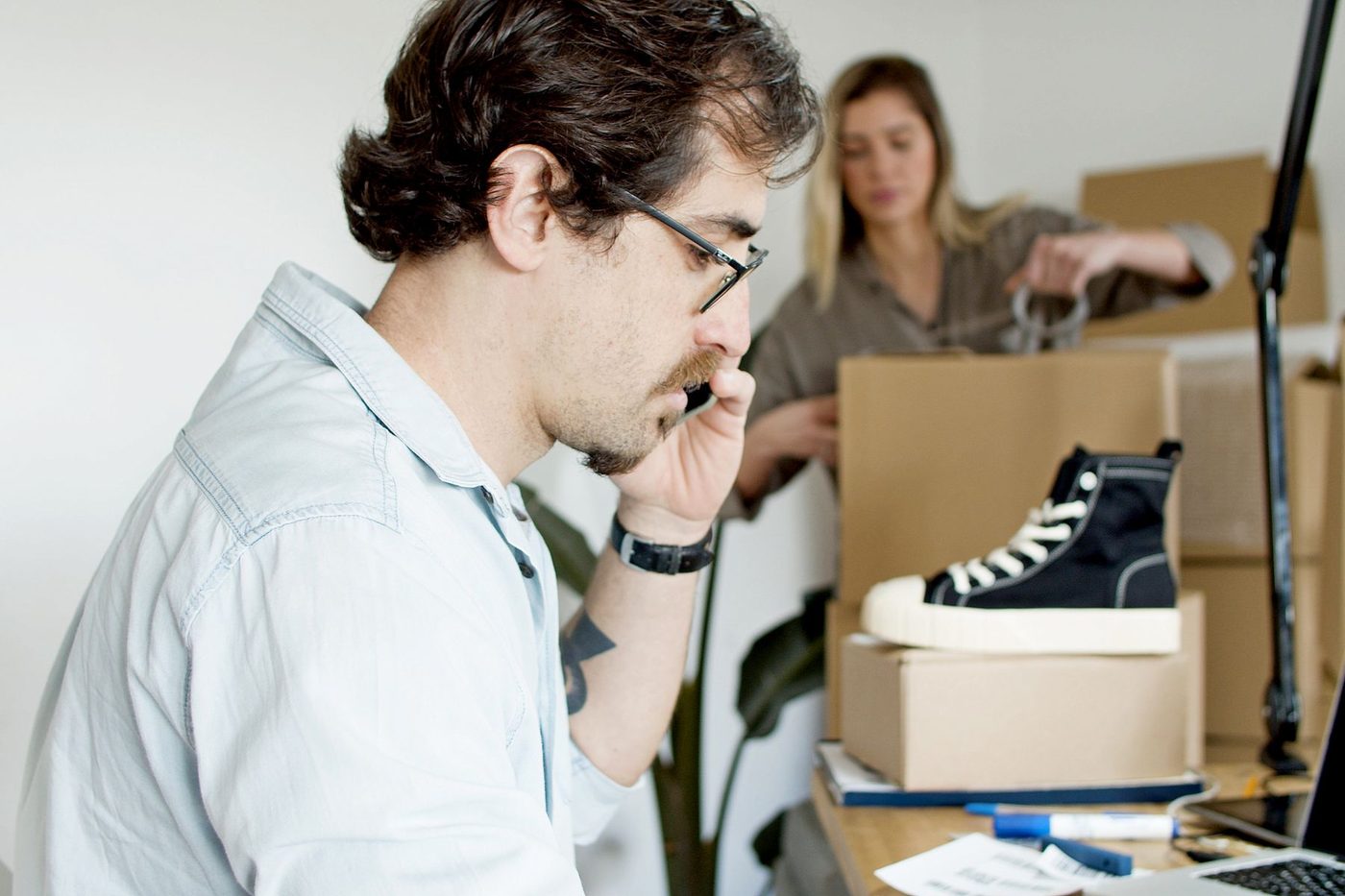 An online shop owner on the phone at his desk while a colleague packs orders behind him